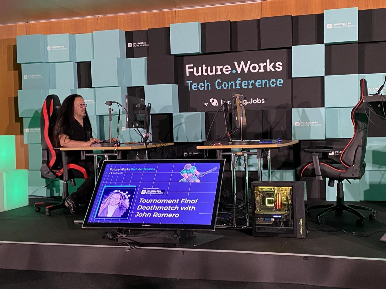 John Romero seated at a computer on the Future.Works Tech Conference stage before a Doom deathmatch